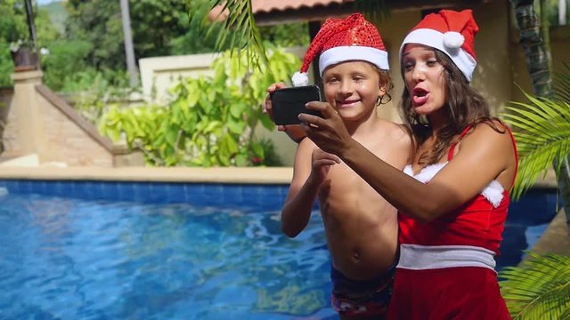 Christmas beautiful mother and child wear christmas hats taking picture self portrait on smartphone together in slow motion near the swimming pool on tropical island of Koh Samui. Thailand. 1920x1080