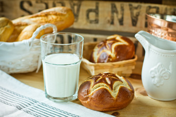 Fresh lye rolls in a wicker basket, a glass of milk, white pitcher on a wood table in a rustic style kitchen interior, copper tableware in the background, cozy atmosphere
