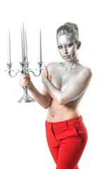 Young topless woman with evening hairstyle and upper body part and hair painted with silver color wearing red trousers posing hiding breast with one hand and keeping candlestick with five candles
