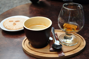 Dark ceramic cup of coffee, glass of cognac and italian almond cantuccini cookie on a white plate on an old wooden table at a cafe.