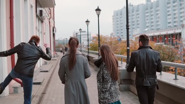 Two trendy girls and two stylish men cheerfully walk in the city, jump and have fun. Slow mo, steadicam shot, back view