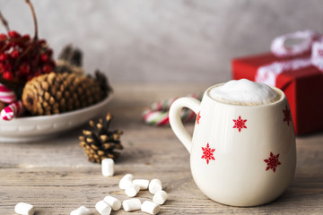 Obraz na płótnie Canvas white cup with winter pattern with milky cappuccino on wooden light desk with christmas and new year decoration like candies, cones, berries and gifts wrapped in red paper