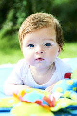 Newborn girl with blue eyes in the beautiful park outdoors, lies on a blue blanket with some toys and looking around.