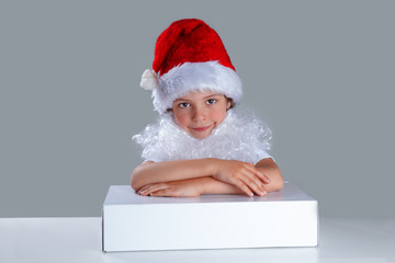 Little Santa Claus put his hands on the white box. He is sitting at a white table. He looks into the camera. Gray background. Close-up.