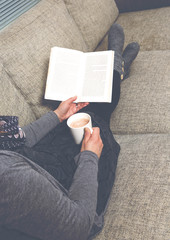 Woman is reading a book on a sofa. Cozy outfit. She is wearing wool socks. Hot drink on a mug. Image has a vintage effect.