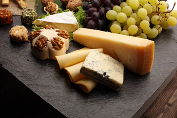 Assorted cheeses on round wooden board plate Camembert cheese, cheese grated bark of oak, hard cheese slices, walnuts, grapes, crackers, bread, thyme, dark black wood background