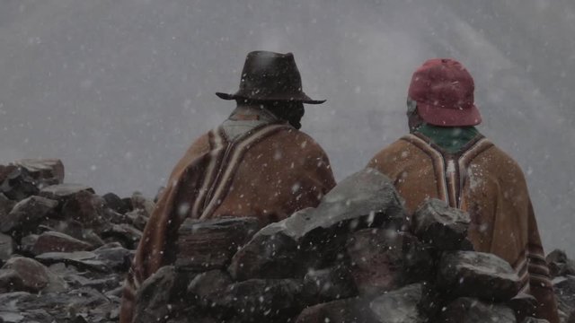 Peruvian muleteers seated on the rocks during a snowfall near the Nevado Pastoruri waiting for the tourists. Slow motion