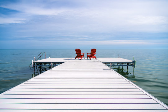 Red Adirondack Or Muskoka Chairs On The Dock Overlooking The Water At The Cottage