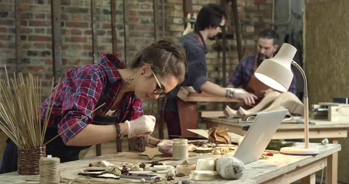 We see a busy workshop. The hipster girl on the foreground is painting wooden toys, and two men, young and middle-aged are making the cabriole table leg/Busy Hipster Workshop