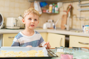 Little blond kid plays with tins for making ginger biscuits, sitting at the kitchen table with tray of raw blanks, ready for baking.