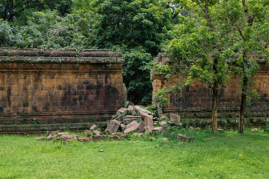 Ancient stone wall with gap from a break through, some loose stone blocks from the breach are visible in front. The wall is located in the antique ruins of Angkor.
