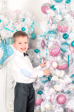 boy blonde in white shirt and pants, standing near the Christmas tree. New Year. Christmas