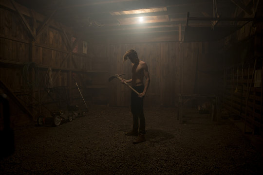 Bare chested man standing in dark barn holding  pickaxe