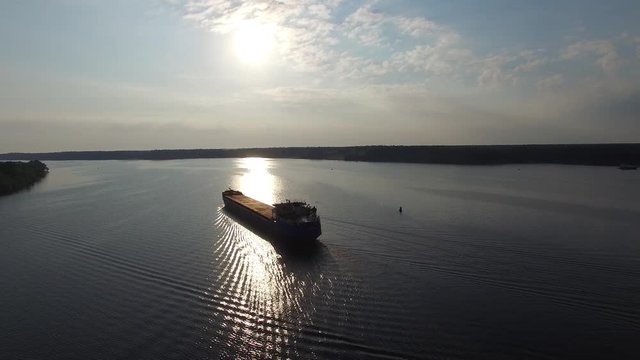 Dry cargo transport ship sailing on a wide river. Sun reflected in calm water. The camera moves in the air away from the ship. The island in the middle of the river. Freight on the river. Aerial view.