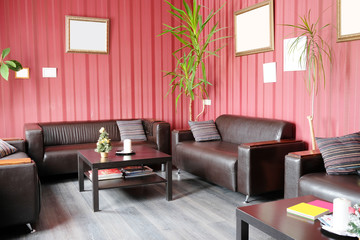 Interior of an empty visitor room in business center with skin furniture in it, green flowers, pictures on a wall with red wall-paper