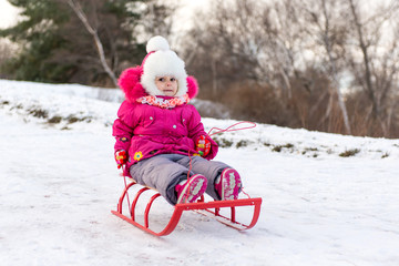 Fototapeta na wymiar The child, a little girl riding on a sled with snow slides. Winter fun for children.
