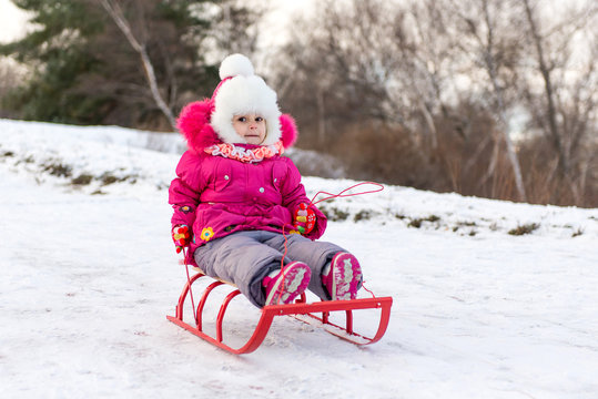 The child, a little girl riding on a sled with snow slides. Winter fun for children.