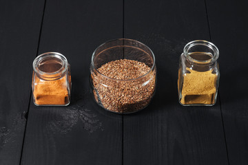 Ground spices in bottles with grain flax in glass plate on black wooden background.