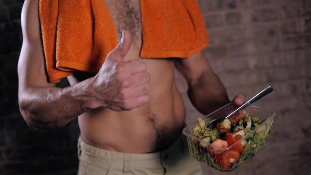 Fitness bodybuilder man holding a plastic bowl and eating fresh salad, on brick background