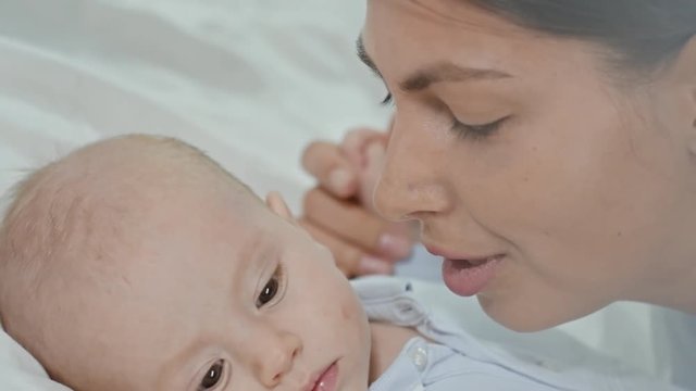 Mother talking to little baby boy lying on bed, then rubbing off lipstick stains of his cheek 