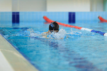 Young woman in goggles and cap swimming breaststroke stroke style in the blue water indoor race pool.