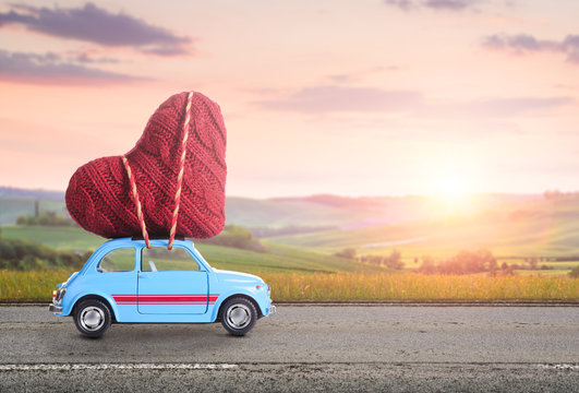Blue retro toy car delivering heart for Valentine's day against blurred rural Tuscany sunset landscape