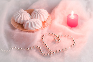 Fototapeta na wymiar White airy zephyrs in the dish on the soft cloth with a candle and decorative, small balls chain in heart form. Very beautiful surprise for someone in special moment.. Romance. Focus on the zephyrs.
