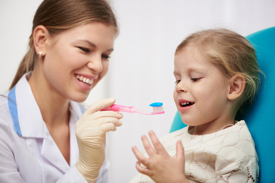 Female hygienist teaching little girl to brush teeth in dental clinic. Dental care concept.