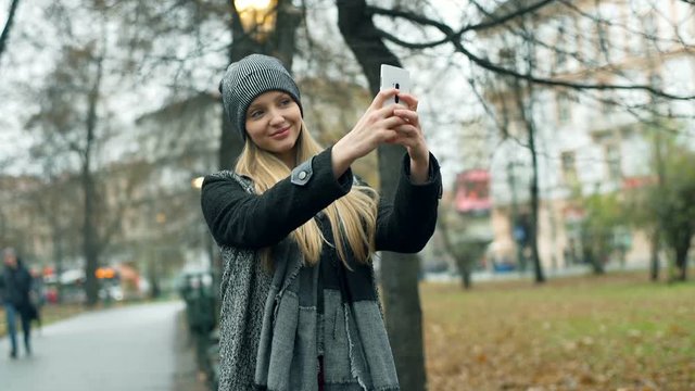 Pretty girl standing in the autumnal park and doing selfies on smartphone, steadycam shot
