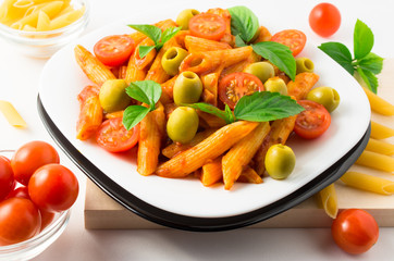 White plate with cooked penne pasta, green olives and red cherry