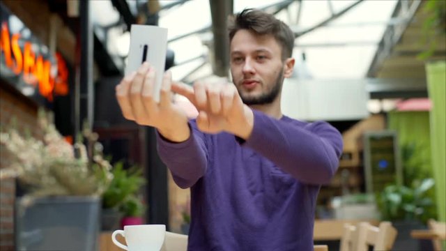 Young man sitting in the cafe and doing selfies on smartphone, steadycam shot
