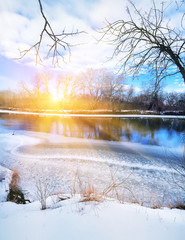 half-frozen river and the coast in the snow in a  in a frosty winter morning. The sun rises from behind the trees. Beautiful sky with clouds.
