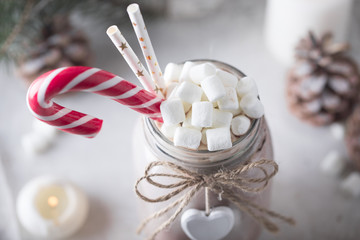 Cocoa with marshmallow, candy cane and straws in the glass jar decorated with little white heart on the table with winter decor for holidays. Close up
