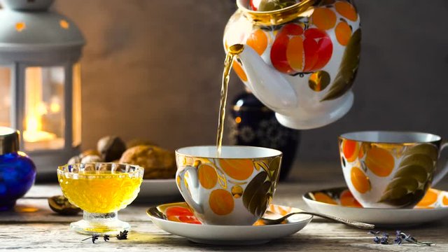 tea time, tea is pouring from the teapot in old fashioned cups of orange and gold colors on wooden desk with candle and honey 