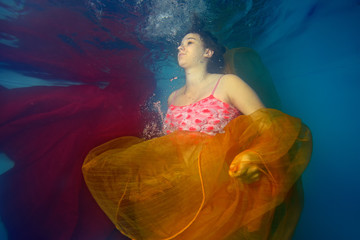 Fototapeta na wymiar Pregnant girl swims underwater and plays with red and yellow cloth on light blue background. Portrait. The view from under the water. Horizontal orientation
