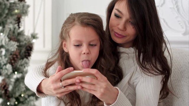 Cheerful young mother and her daughter making comic xmas selfies and showing tongues