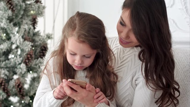 Young mother teaching her daughter how to use smartphone in Christmas decorated room
