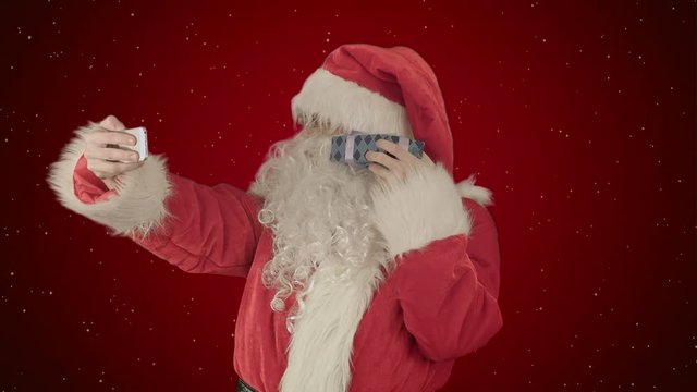 Santa Claus holding a big present  doing a selfie on smartphone on red background with snow