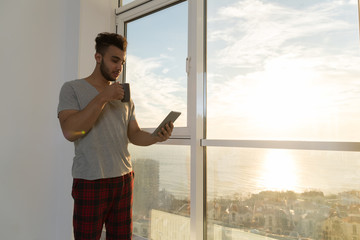 Hispanic Man Using Cell Smart Phone Modern Apartment Big Panoramic Window Sea View, Young Guy Morning Hold Cup Chatting Online Home Interior