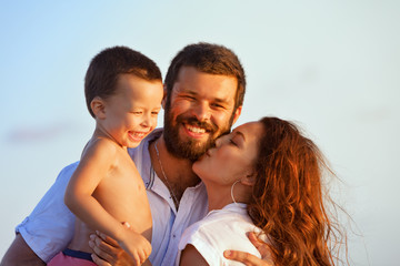 Face portrait of hugging beautiful family - happy mother, father holding baby son on hands walk with fun on sunset beach. Active parents, people outdoor activity on tropical summer holiday with child
