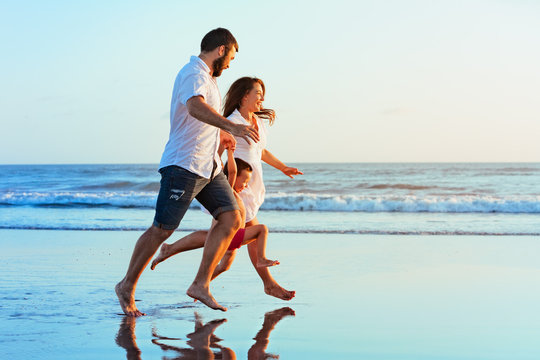 Happy family - father, mother, baby son hold hands, run together with splashes by water pool along sunset sea surf on black sand beach. Travel, active lifestyle, parents with child on summer vacation.