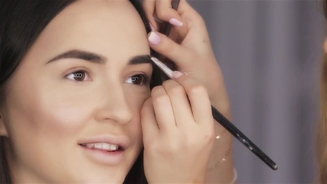 Make up artist paints eyebrow of beautiful brunette young woman close-up