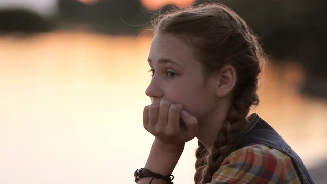 Sad teen girl (14-15) resting chin in hand thinking outdoor. Portrait of pensive teenager looking away sitting by calm lake at sunset.