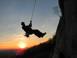 Kletterer hängt am Seil vor Sonnenuntergang, oberhalb Gleitschirmflieger