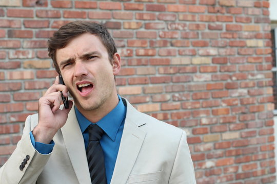 Guy on the phone with headache. Upset unhappy male talking on phone isolated on brick wall background. Negative human emotion face expression feeling life reaction. Cellular mobile radiation concept