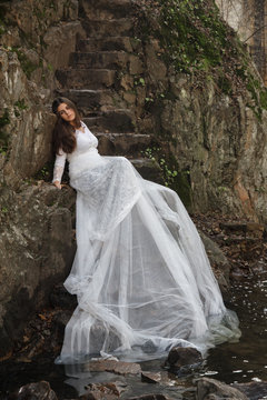 Young woman with blue eyes dressed bride sitting on a stone stairs in the countryside near a river just before marriage. 