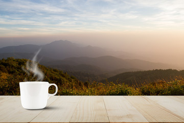 Hot coffee cup with steam on vintage wooden table top on blurred meadow and foggy mountain background during sunrise