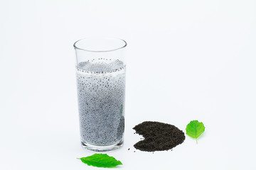 Glass of hairy basil seeds with water and dried seeds on floor with green leaves on white background