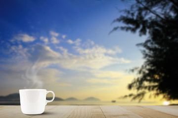 Hot coffee cup with steam on wooden table top on blurred blue sky and beach during sunrise