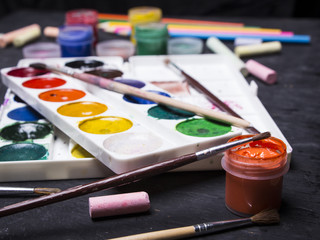 Fototapeta premium Bright multi-colored paint, pastels, crayons, pencils, brushes on a black wooden background. Set for the artist. Education.